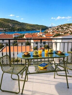 En balkon med et bord dækket til morgenmad, med udsigt over en kystby med bygninger og en bro.