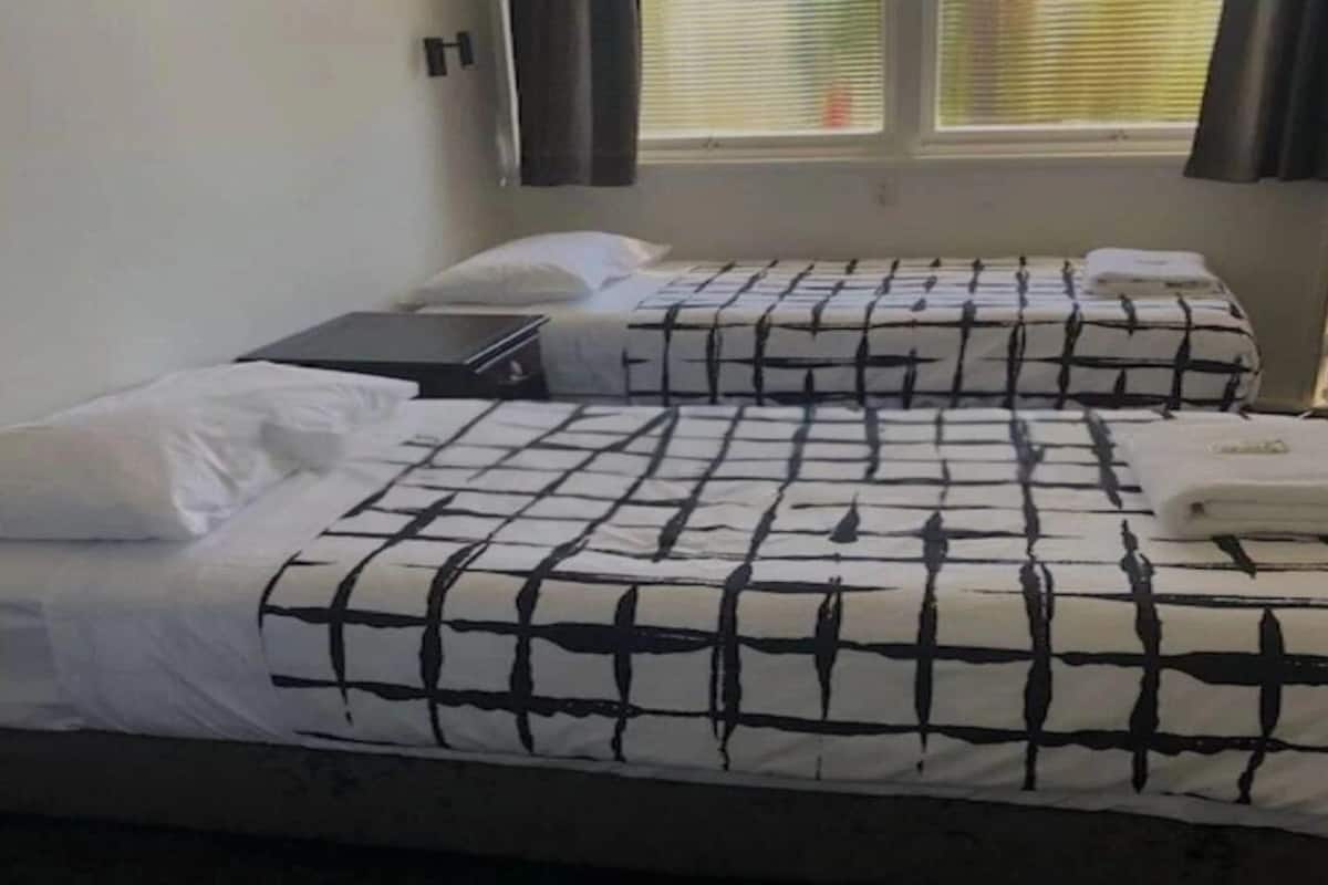 A room with two beds, each with a black and white checkered bedspread and white pillows. There is a window with blinds partially open, allowing natural light to enter.
