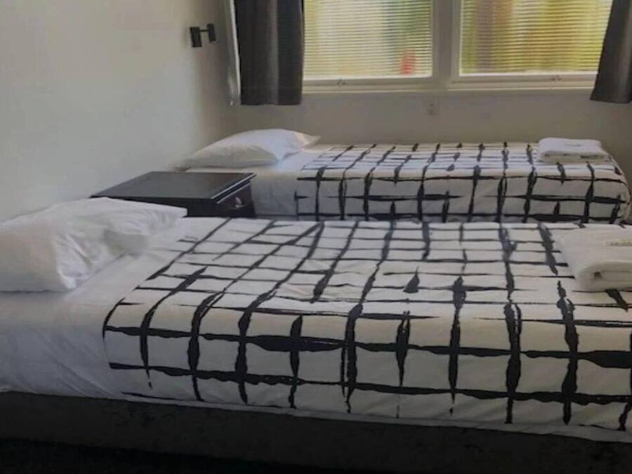 A room with two beds, each with a black and white checkered bedspread and white pillows. There is a window with blinds partially open, allowing natural light to enter.