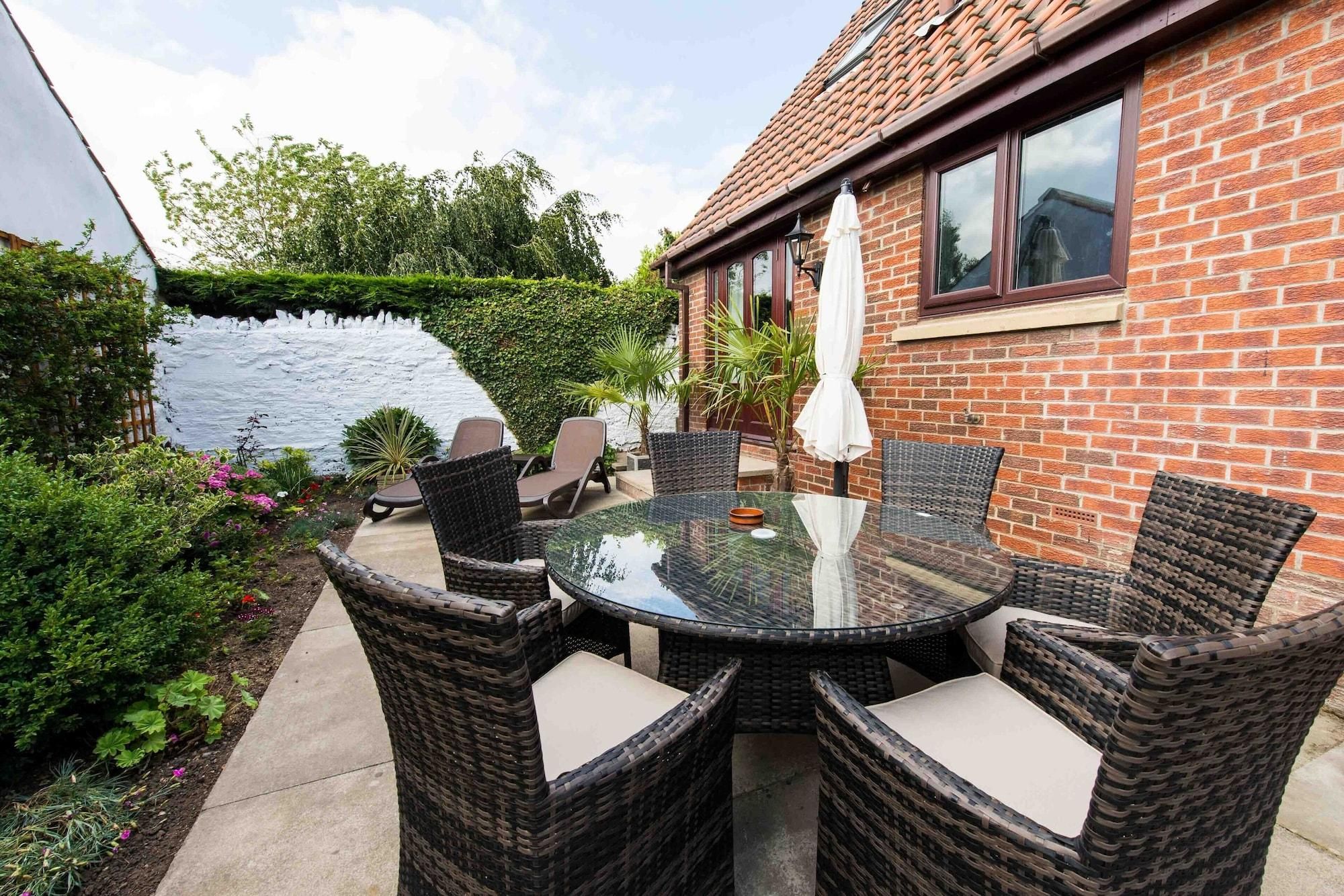 Une terrasse avec des chaises en osier et une table en verre, entourée d’une maison en briques et d’un mur blanc.