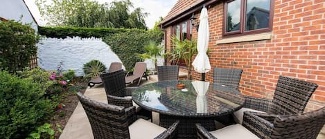 Une terrasse avec des chaises en osier et une table en verre, entourée d’une maison en briques et d’un mur blanc.