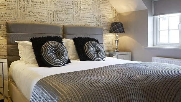 A bedroom with a patterned wallpaper, a bed with a striped bedspread, two black and white pillows, and a bedside lamp. - The Copper Horse Cottages (Scarborough)