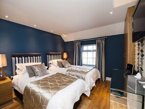 Quarto de hotel com duas camas, uma janela grande com cortinas, piso de madeira e uma escrivaninha com computador.
