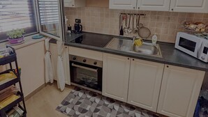 Une cuisine avec des meubles blancs, un plan de travail noir, un four encastré, un four à micro-ondes, une fenêtre avec des stores et un tapis à motifs.