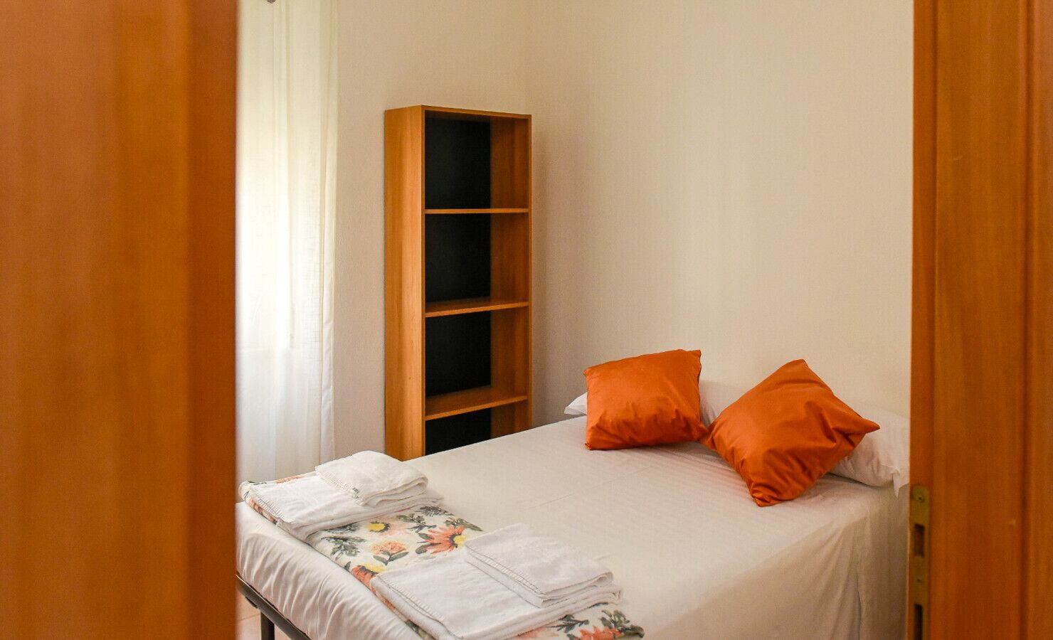 Uma cama de solteiro com colcha floral, duas almofadas laranja e uma estante de madeira contra uma parede lisa.