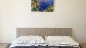 A neatly made bed with a grey quilt and white pillows, two rolled towels with blue ribbons, and a framed picture of a coastal landscape on the wall. - Villa the Scent of Oranges (Eboli)