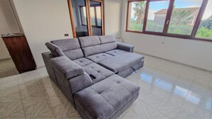 A spacious living room with a grey sectional sofa, a wooden cabinet, and a large window offering a view of greenery. - Villa the Scent of Oranges (Eboli)