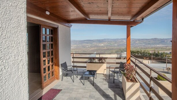 A covered balcony with a view of a valley, featuring a wooden ceiling, a glass door, and outdoor seating.