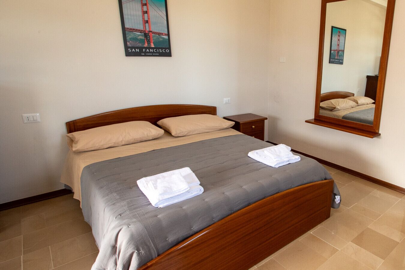 Een opgemaakt bed met een houten hoofdbord, een klein nachtkastje en een spiegel aan de muur.