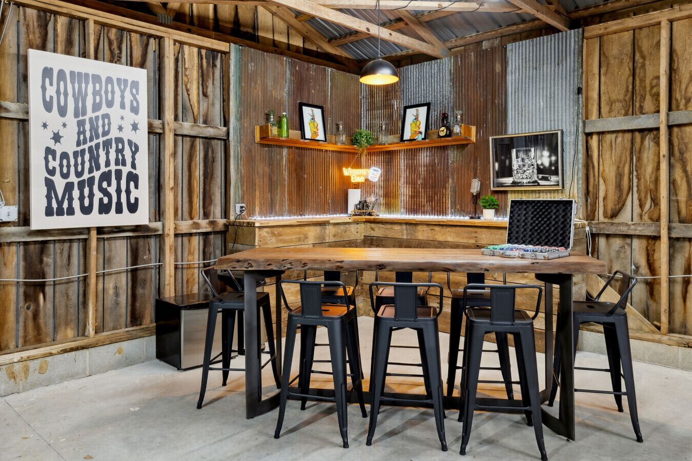 Un rústico espacio de bar con una mesa de madera y sillas negras, un cartel que dice “COWBOYS AND COUNTRY MUSIC” y estanterías con diversos objetos.