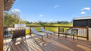 A wooden deck with a barbecue grill, lounge chairs, and a view of a green landscape.