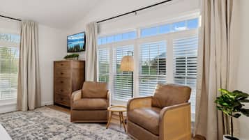 A spacious room with a large window, a sofa, a chair, a small table, a dresser, and a potted plant.