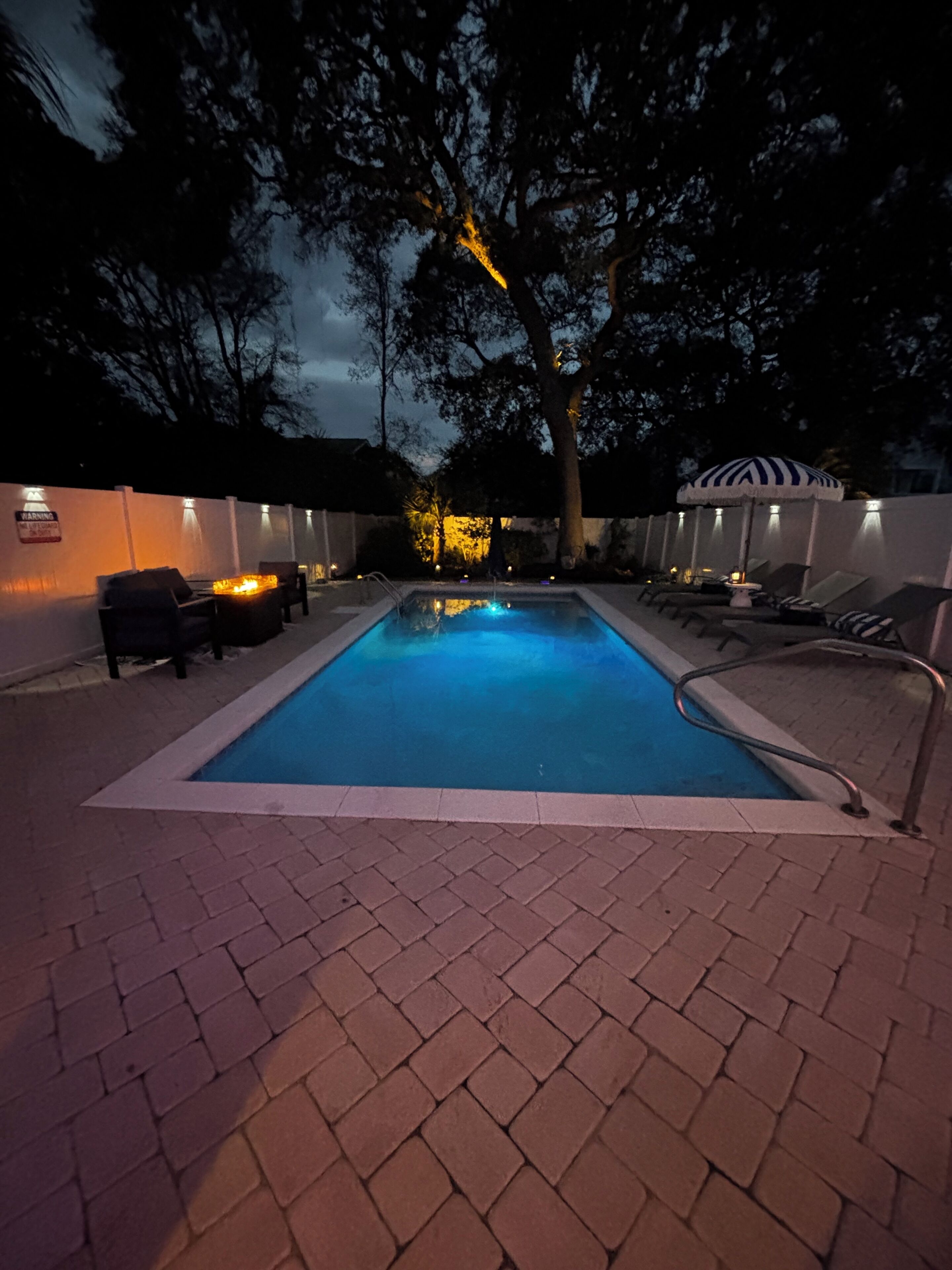 Ein rechteckiger Pool mit einer gefliesten Kante, umgeben von einer Pflasterterrasse. Auf einer Seite befinden sich Liegestühle und ein Tisch mit Sonnenschirm, auf der anderen Seite eine Mauer mit einem 'No Parking'-Schild.