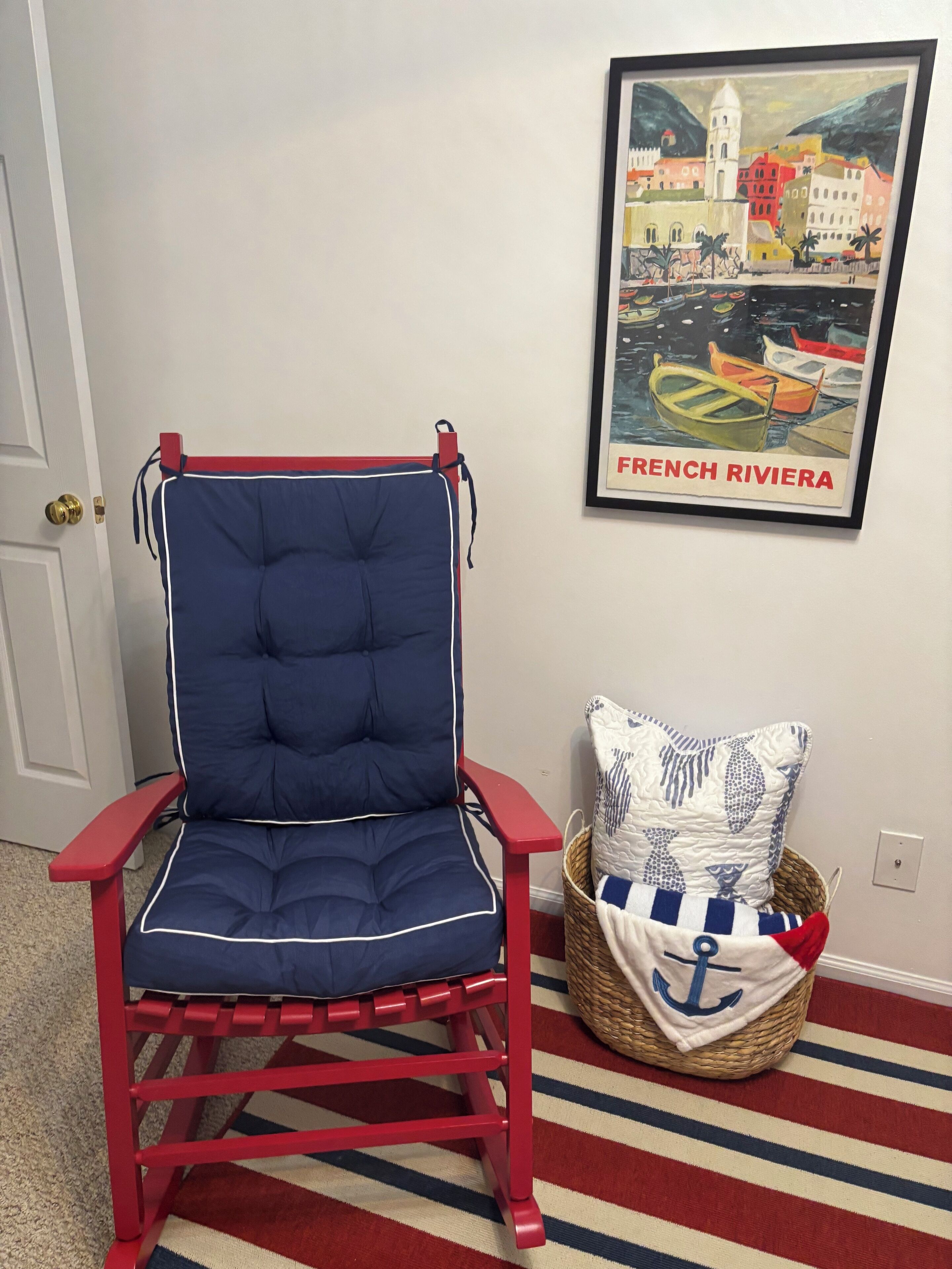 A red and blue chair with a cushion, a framed picture of French Riviera, and a basket with a pillow on a striped rug.