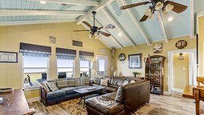A spacious living room with a large sectional sofa, a coffee table, and a fireplace. There are ceiling fans, a bookshelf, and a view of the outdoors through large windows. - Ocean View~Elevator~PetFriendly~Games~Beach Access (Bolivar Peninsula)