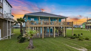 A two-story house with a balcony, surrounded by a well-maintained lawn and palm trees. - Ocean View~Elevator~PetFriendly~Games~Beach Access (Bolivar Peninsula)