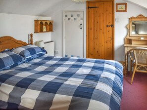 A bedroom with a checkered bedspread, a wooden door, a white bedside table, and a wicker chair. - Rivers View - UK49662 (Braunton)