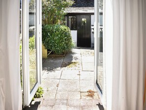 A narrow pathway leading to a building with a black door, surrounded by greenery and a stone-paved area.