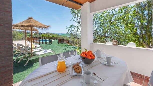 Un coin repas avec une table dressée pour le petit-déjeuner, un parasol en chaume et une vue sur une piscine et un jardin.