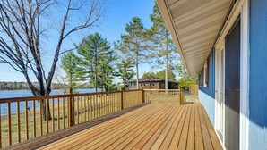 Um deck de madeira com vista para um lago e árvores, uma área coberta com uma janela e um prédio ao fundo.