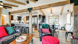 Uma sala de estar ampla com sofá de couro, um banquinho vermelho e uma mesa de centro de madeira. Ao fundo, cozinha com eletrodomésticos modernos e área de refeições com mesa e cadeiras.