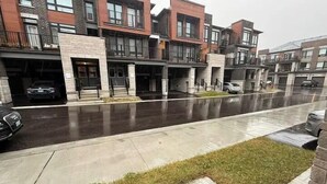 A row of modern townhouses with garage doors, a wet driveway, and a grassy area in the foreground. - Ajax Villa  (Ajax)