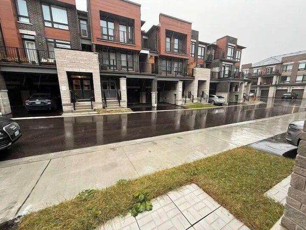 A row of modern townhouses with garage doors, a wet driveway, and a grassy area in the foreground. - Ajax Villa  (Ajax)