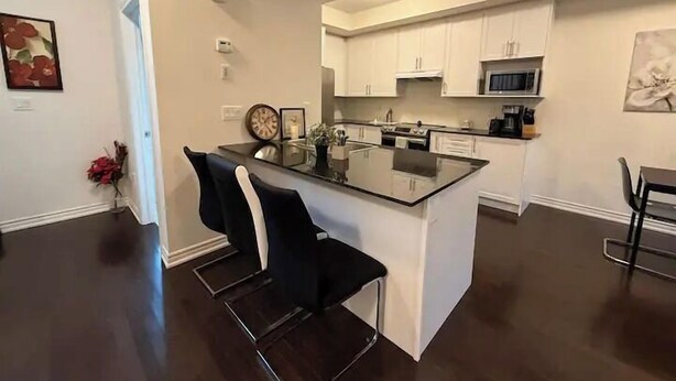 A modern kitchen with white cabinets, a central island, and a dining area with a black chair. - Ajax Villa  (Ajax)