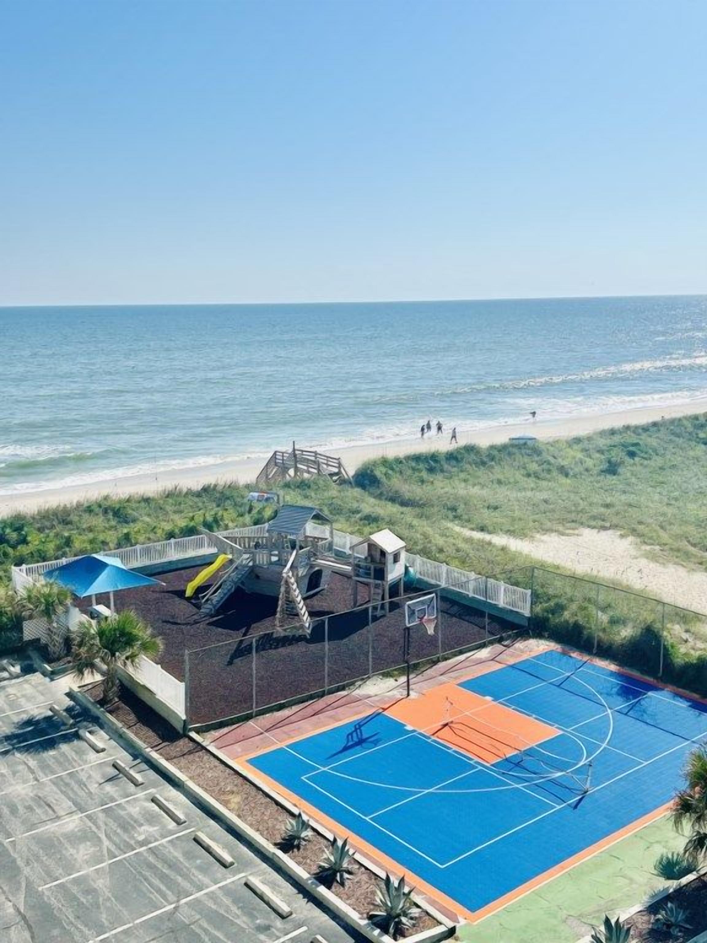 Una cancha de baloncesto frente a la playa, con un parque infantil, un estacionamiento y vistas al océano.