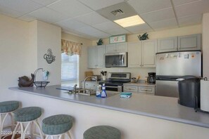 A kitchen with a central island, bar stools, a microwave, a refrigerator, and cabinets. - Sunrise & Serenity - St. Regis Condo #3001 (N. Topsail Beach)