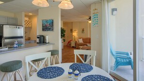 Una cocina con refrigerador, estufa y lavadero. Hay una mesa de comedor con sillas y manteles individuales azules. Un cuadro está en la pared y se ve una ventana con cortinas.
