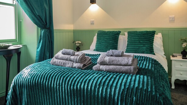 A neatly made bed with green and white bedding, two wall-mounted lights, a window with curtains, and a small table with a vase of flowers. - 2 bedroom accommodation in Staithes (Staithes)