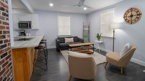 A modern living room with a grey sofa, a coffee table, and two armchairs. There is a kitchen area with a microwave and a coffee machine. A wooden bar counter with stools is visible. - The Little Lake House-Cozy 2Bdrm (Midland)