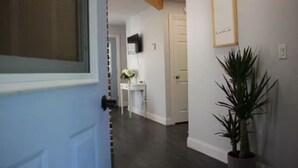 A hallway with a white door, a potted plant, a white wall with a framed picture, and a white wall with a mounted television. - The Little Lake House-Cozy 2Bdrm (Midland)