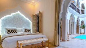 Slaapkamer met een groot bed, een houten bankje, een decoratief hoofdeinde, en uitzicht op een zwembad en binnentuin.