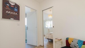 A room with a bed, a small table with books, and a colorful cushion. There is a door leading to another room with a bed and a window.