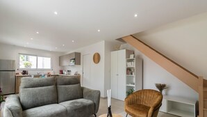 A modern living room with a grey sofa and a brown armchair, a kitchen in the background, and a staircase leading to another level.