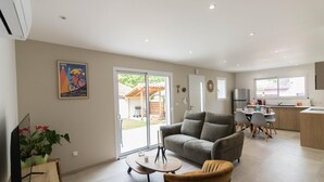 A modern living room with a grey sofa, a small round table, and a brown armchair. There is a dining area with a table and chairs, and a kitchen with a refrigerator and sink.