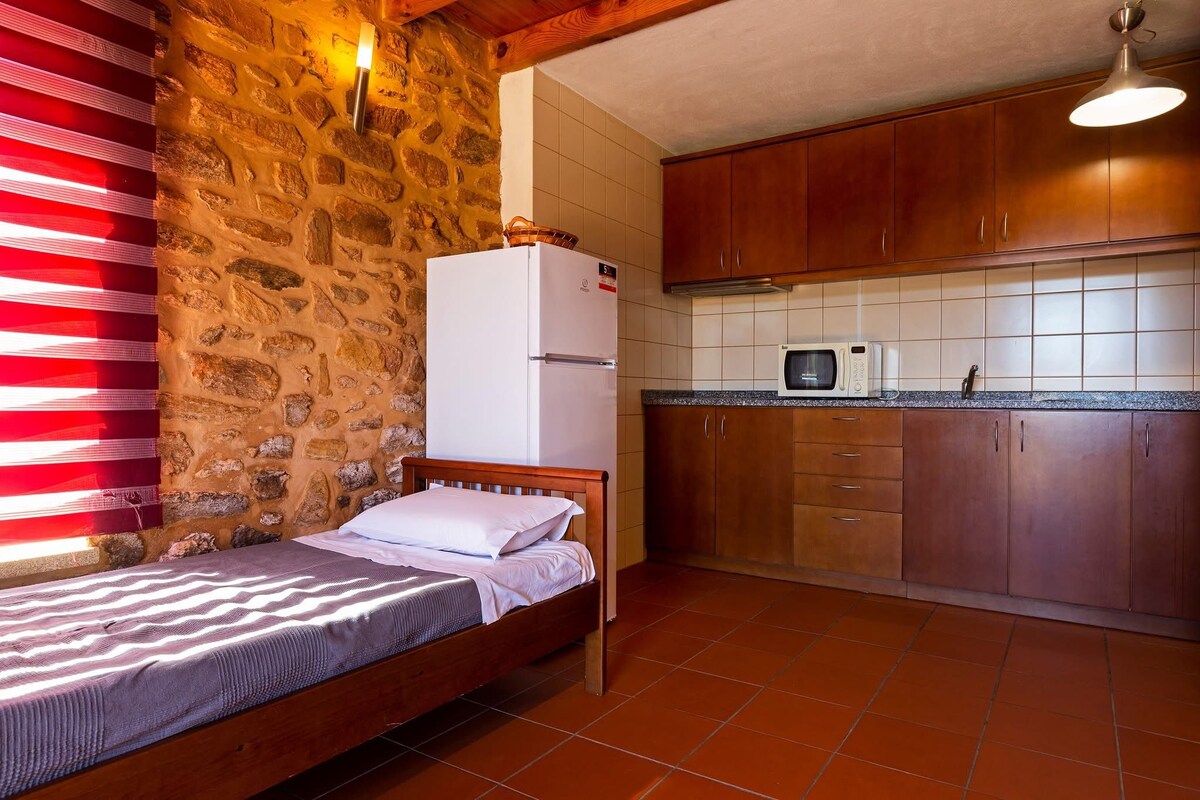 A compact bedroom with a single bed, a stone wall, and a wooden ceiling. Adjacent is a kitchen with wooden cabinets, a microwave, and a refrigerator.
