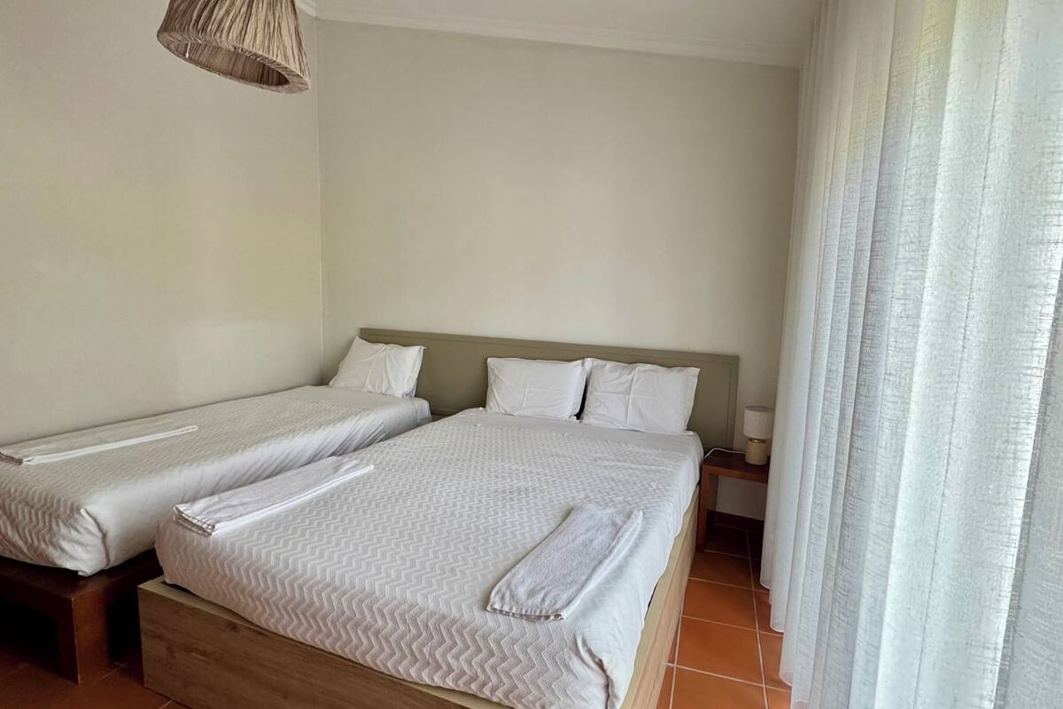Une chambre d’hôtel avec deux lits simples, une tête de lit en bois, une table de chevet avec une lampe et une fenêtre avec des rideaux légers.