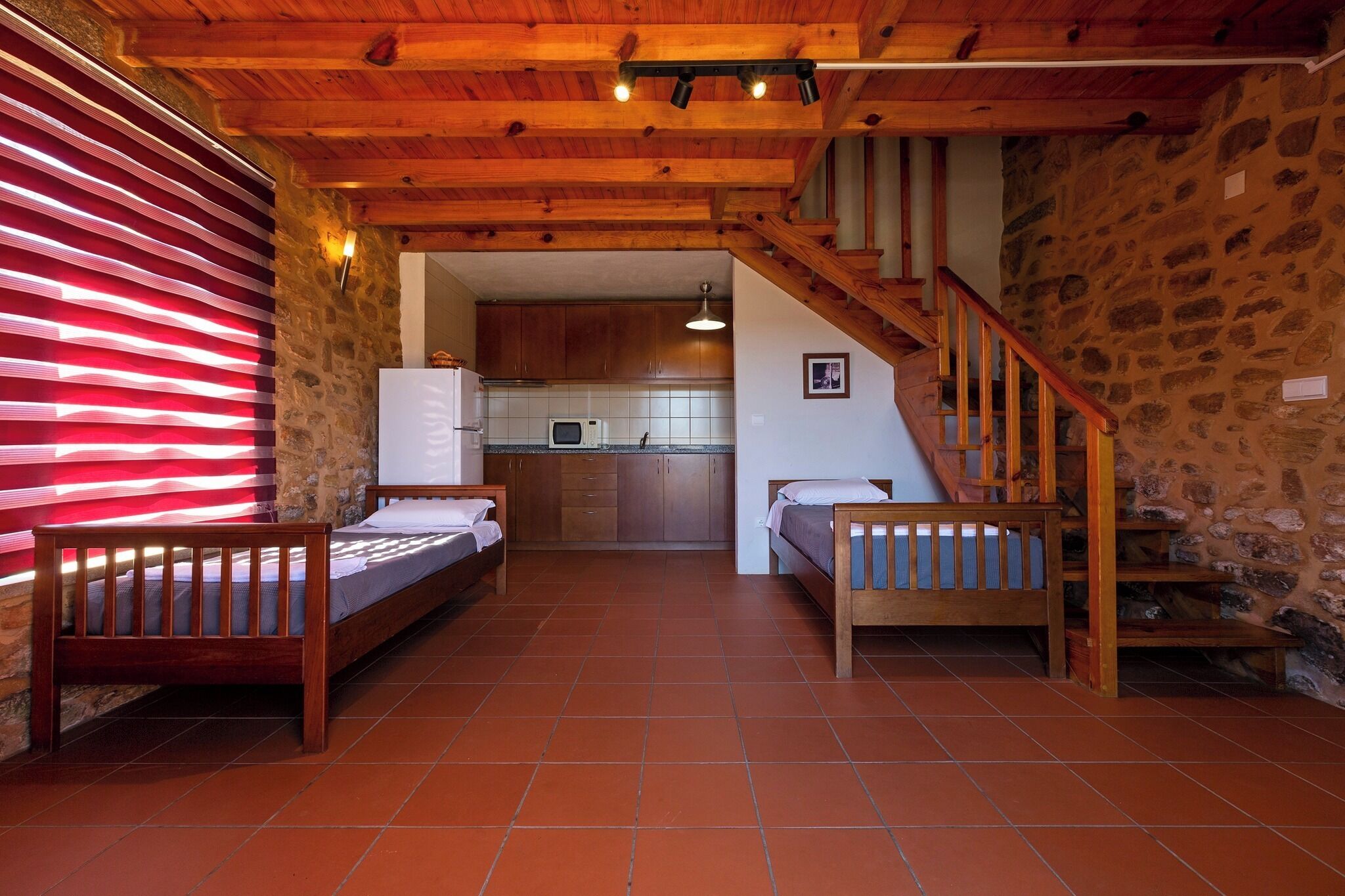 A cozy room with two wooden beds, a stone wall, a kitchen in the background, and a wooden staircase.