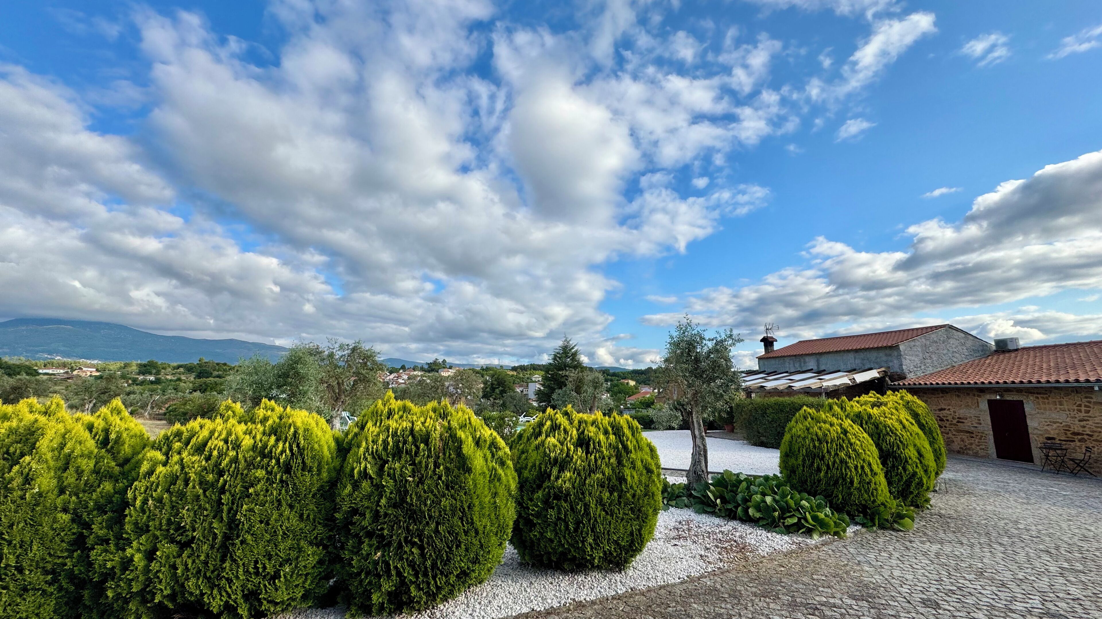 Un patio con setos bien cuidados, un edificio de piedra con tejado de baldosas y un paisaje montañoso al fondo.