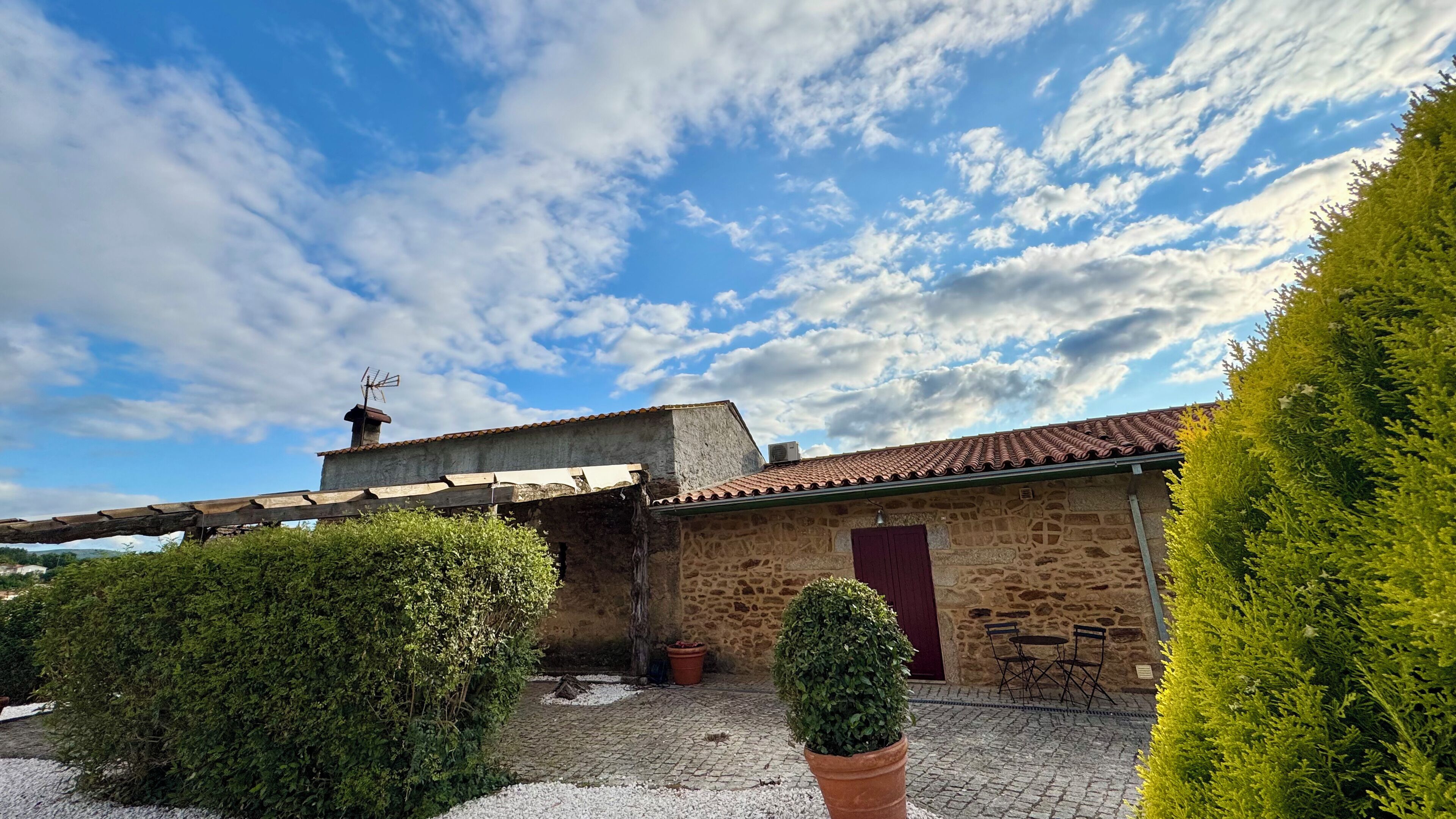 A stone building with a tiled roof, a wooden pergola, and a small garden area with potted plants.
