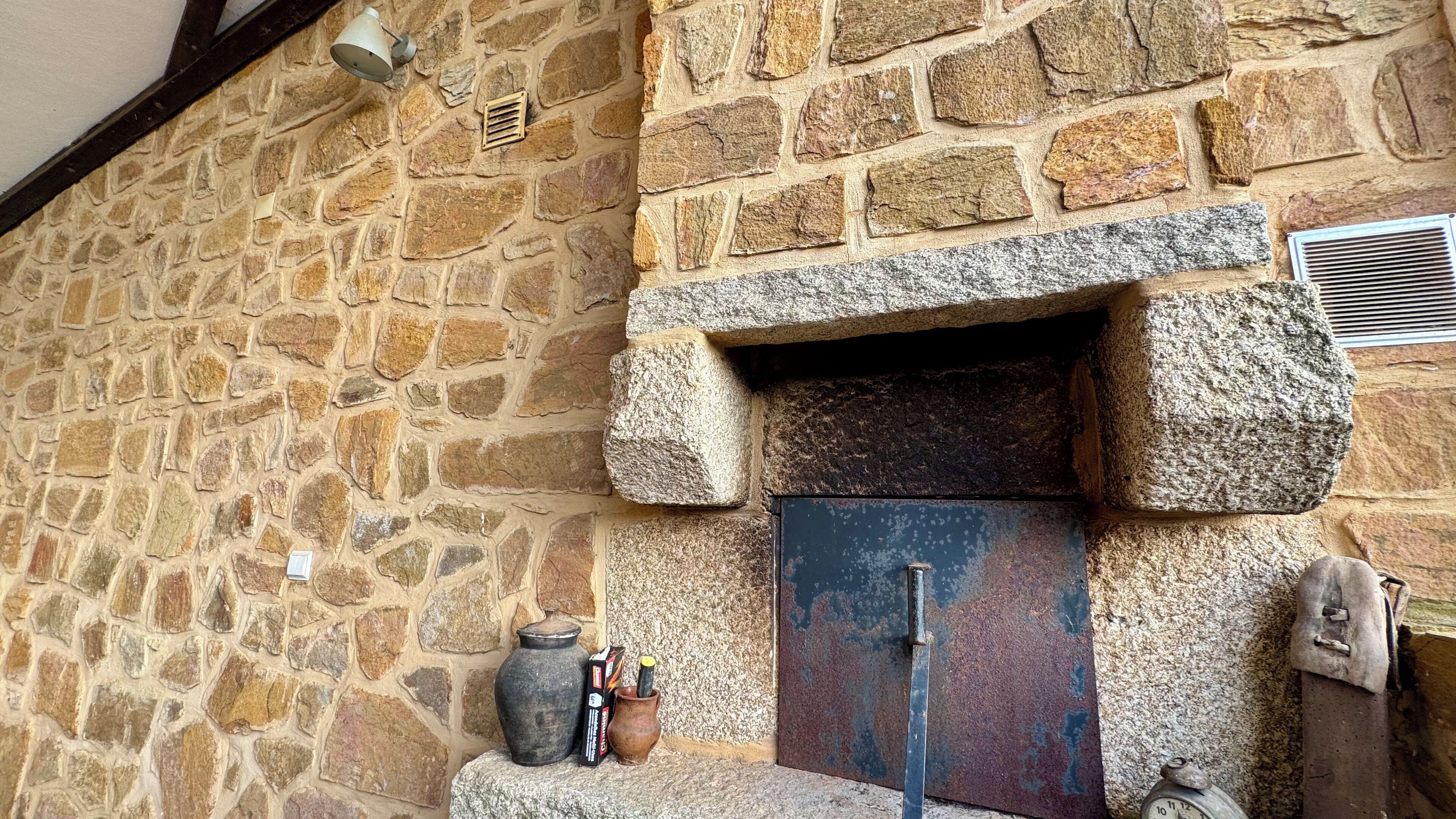 Un muro in pietra con un rustico camino in pietra, una porta in metallo e un piccolo ripiano con un vaso e una scatola.