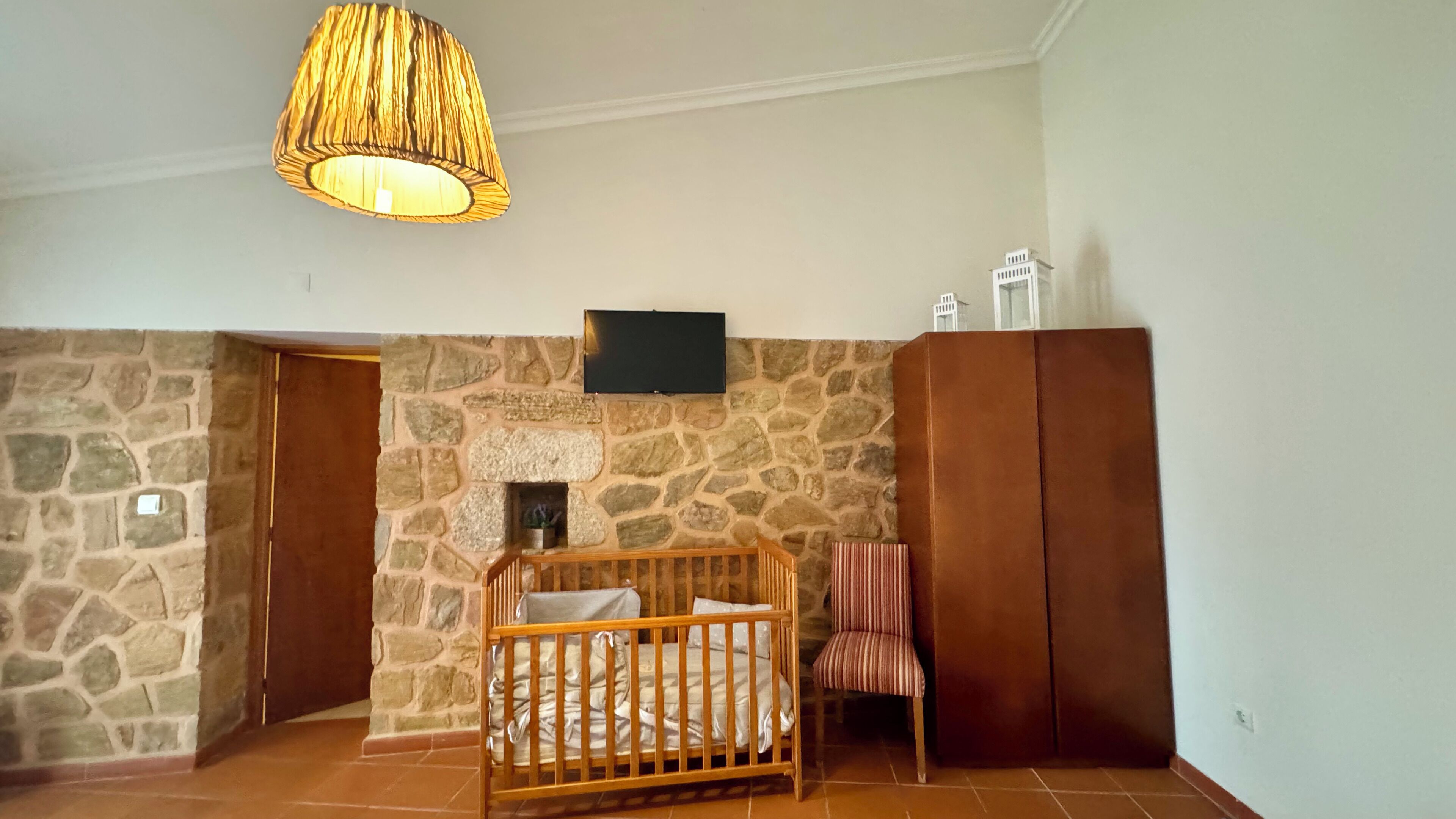 Una habitación con pared de piedra, una cuna de madera, un televisor montado en la pared y un armario.