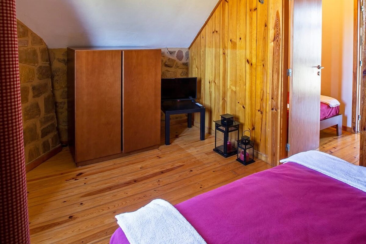 A bedroom with a wooden ceiling, a bed with a pink bedspread, a wooden wardrobe, and a stone wall.