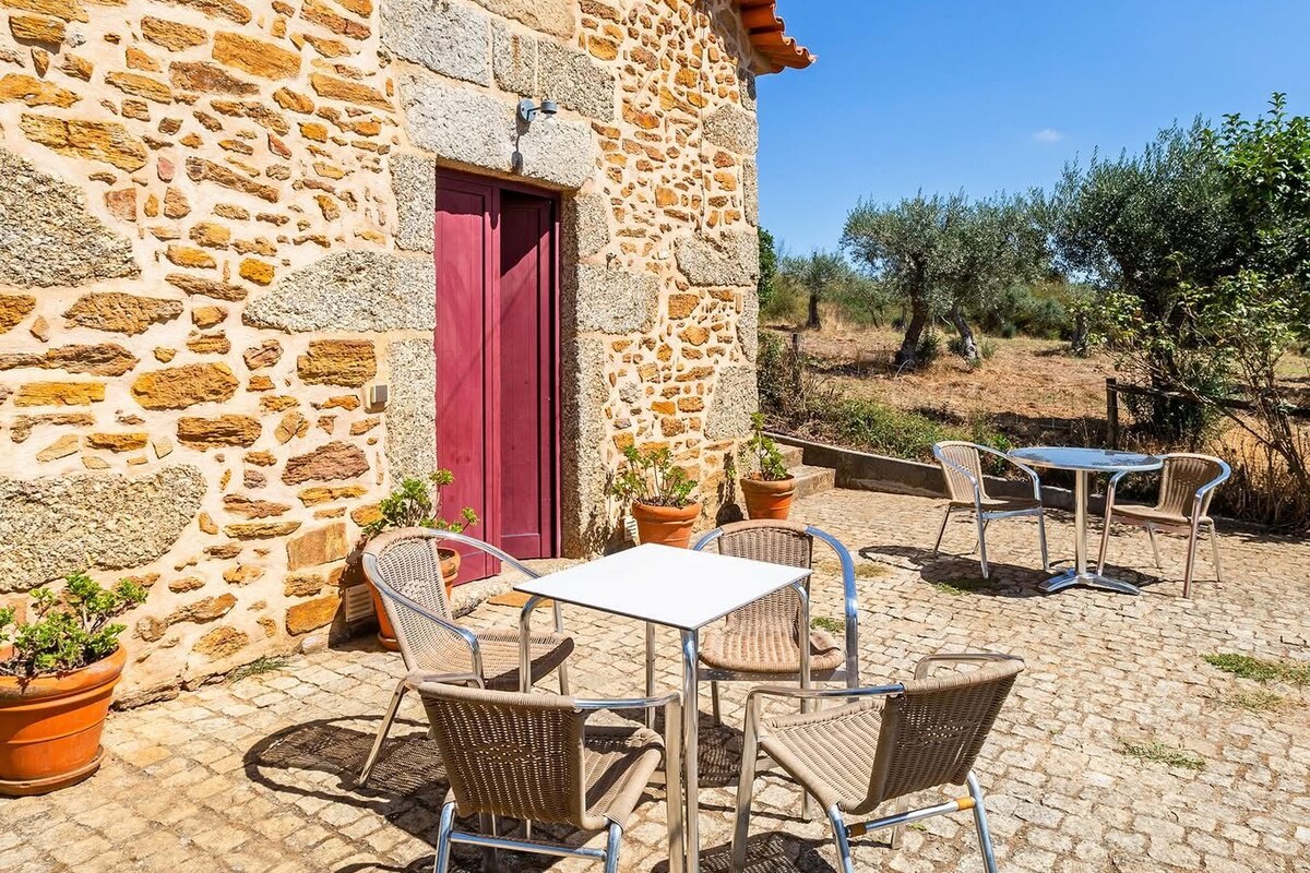 Un bâtiment en pierre avec une porte rouge et une fenêtre, des chaises en osier à l’extérieur et une table sur une terrasse pavée.