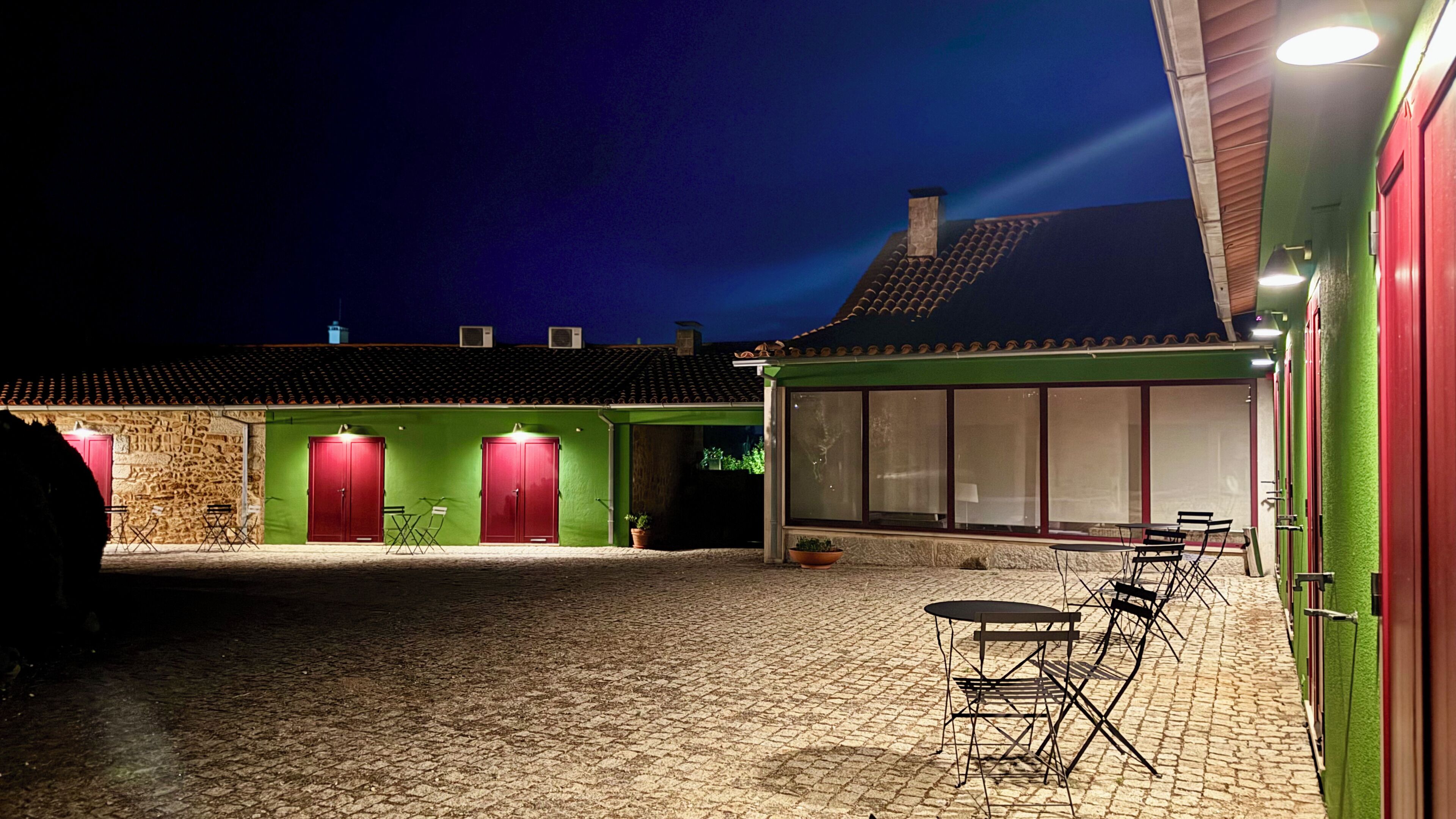 Un patio con suelo de adoquines, un edificio verde con puertas rojas y una zona de estar con mesas y sillas.