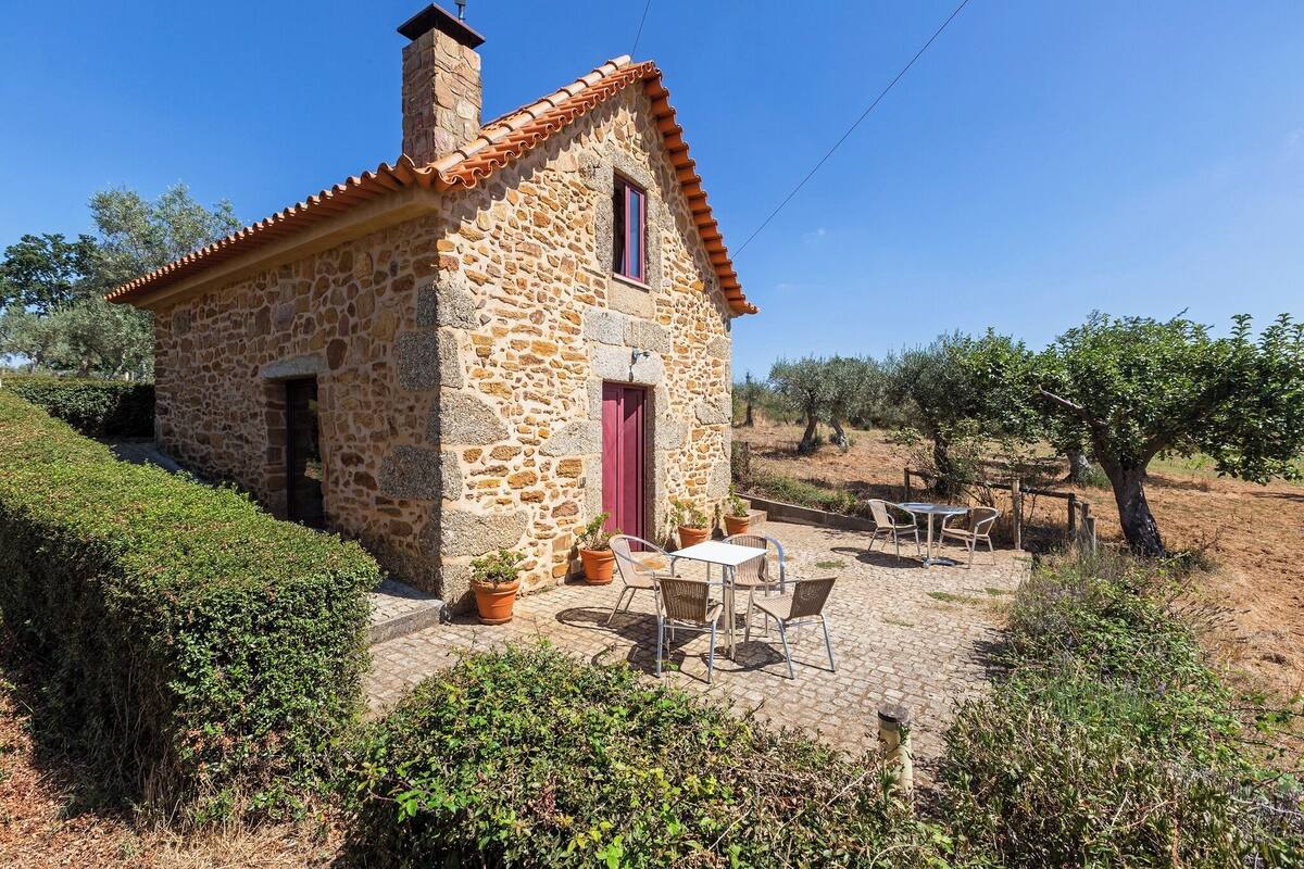Une maison en pierre avec une porte rouge et une petite fenêtre, entourée de plantes en pot et d’une table avec des chaises.