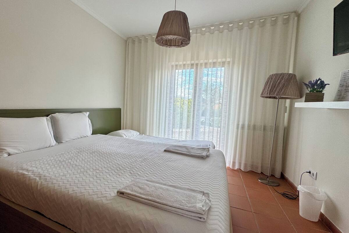 Un lit bien fait, avec du linge de lit blanc et une serviette pliée, une lampe de chevet et une fenêtre ornée de rideaux légers.